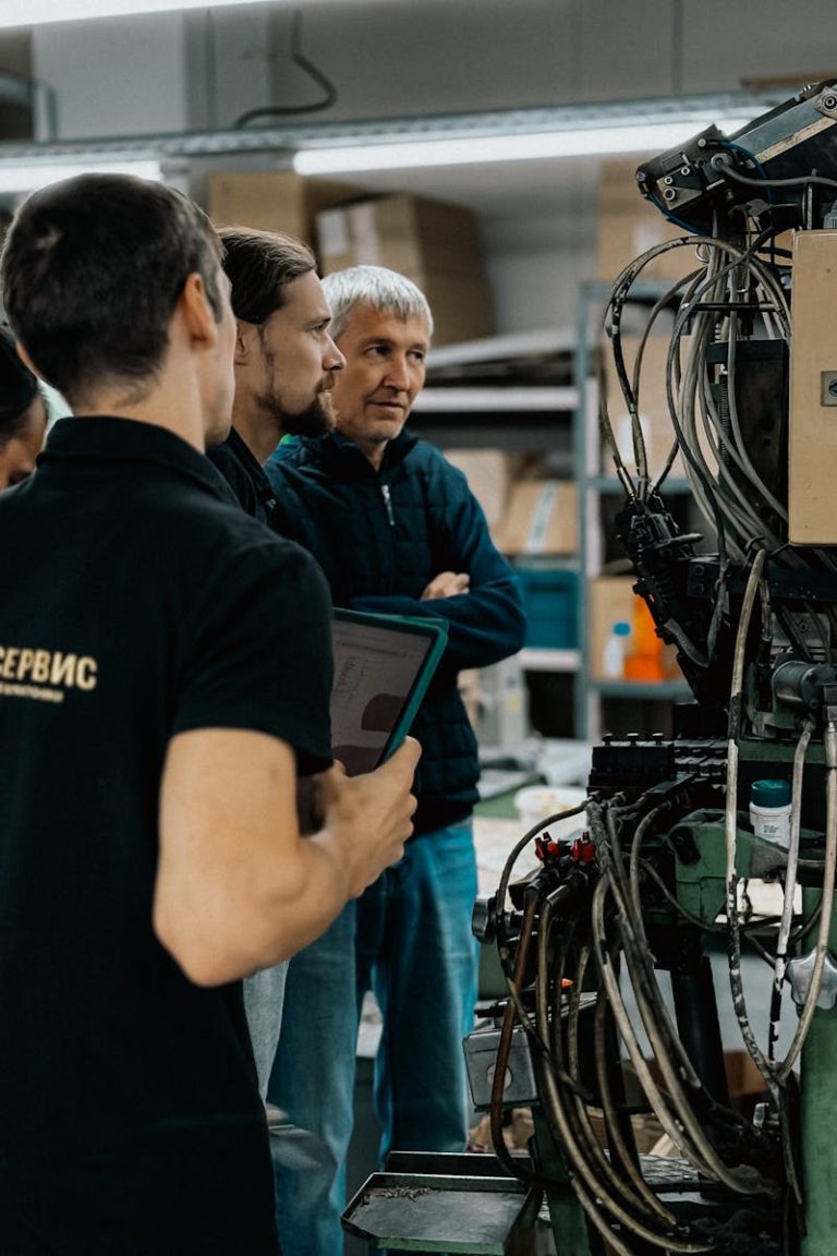 Three adults engaged in a discussion around complex machinery in a factory environment.