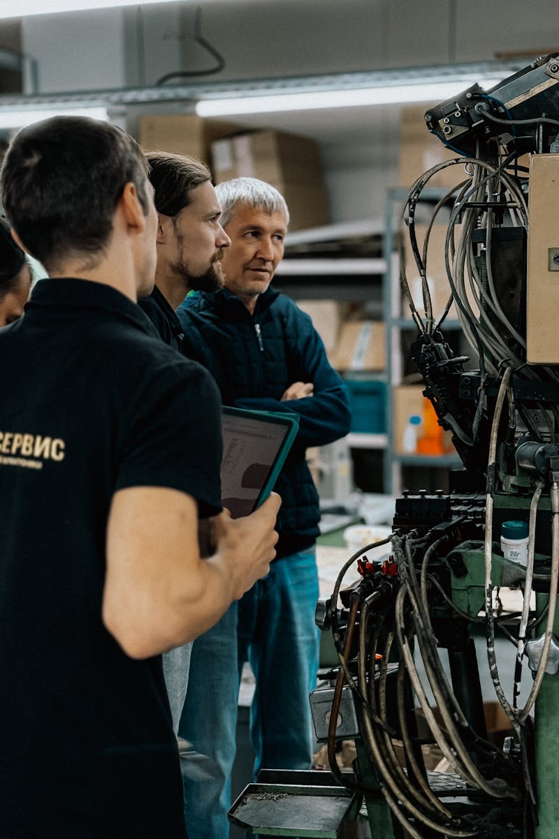 Three adults engaged in a discussion around complex machinery in a factory environment.